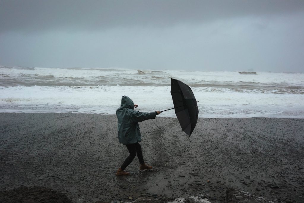 Full body of anonymous person in hood standing in wind with umbrella on sandy shore near stormy sea in rainy weather