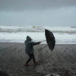 Full body of anonymous person in hood standing in wind with umbrella on sandy shore near stormy sea in rainy weather